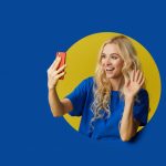 image-of-young-happy-woman-standing-over-a-blue-wall-woman-peep-out-of-a-hole-in-the-wall-looking-to-the-side-to-take-a-selfie-on-the-mobile-phone