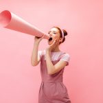 cheerful-woman-with-buns-screams-into-mouthpiece-woman-in-overalls-posing-on-pink-background